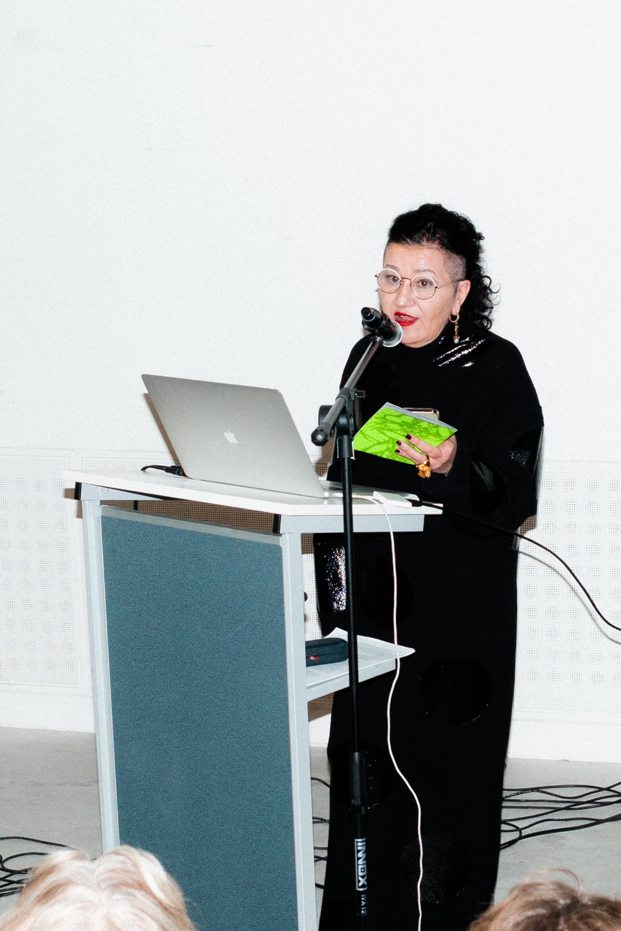 The image shows a woman standing in front of a laptop, presenting at an event. She is wearing dark clothes and wears red lipstick.