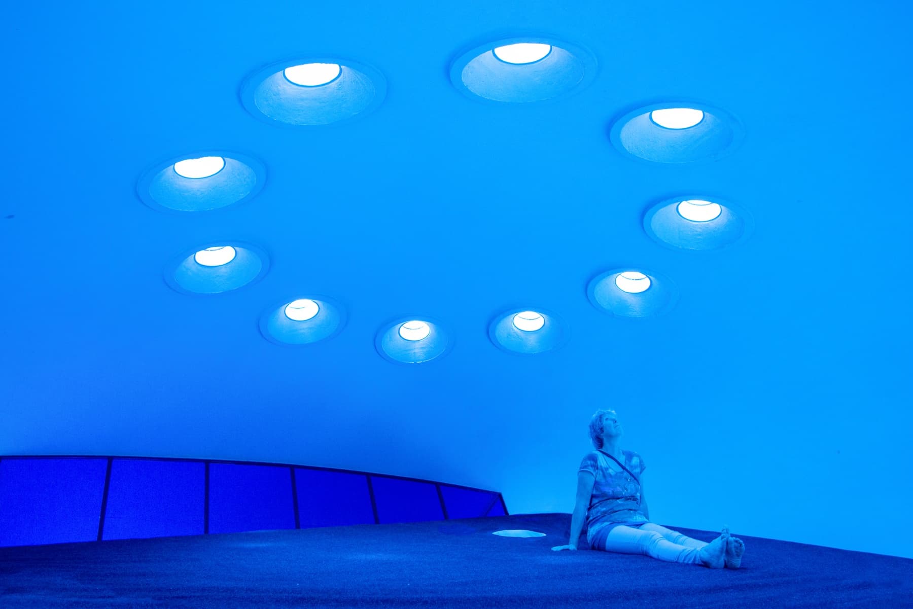 The image shows a room lit blue. A person is sitting on the floor and looking up.