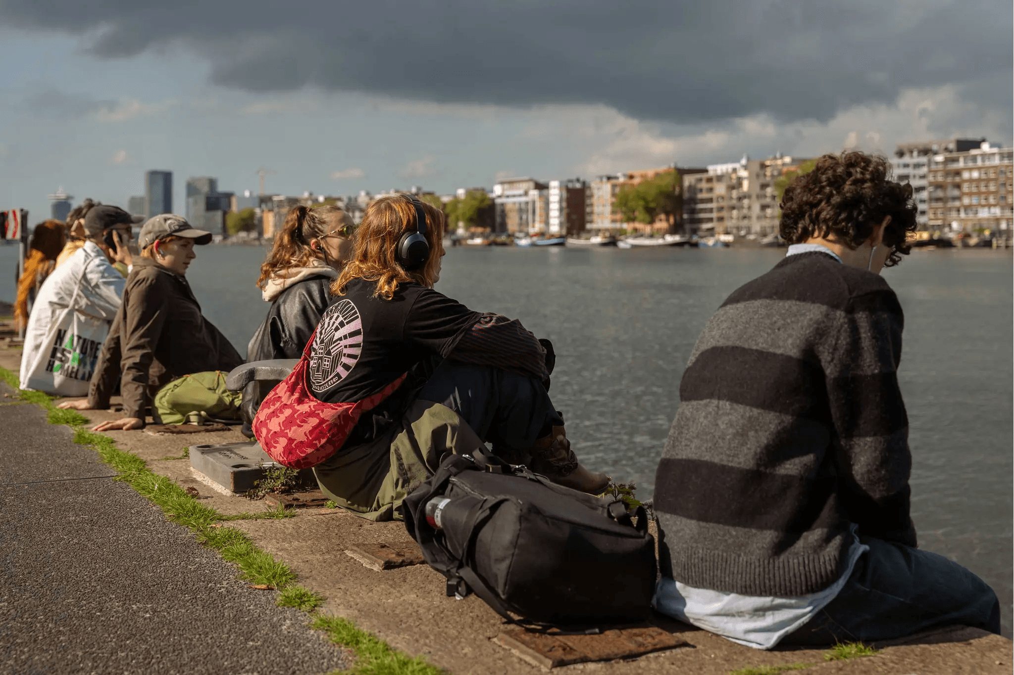 The image shows a group of people wearing headsets sitting on the edge of a river, listening to something. The sky is overcast.