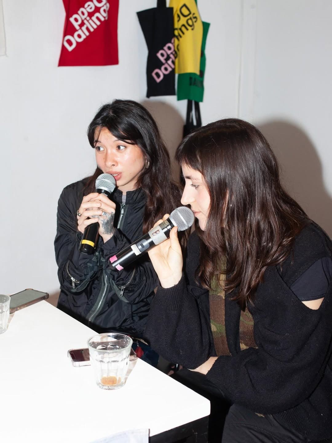 The image shows two femme-presenting people with long dark hair sitting at a table and speaking into microphones.