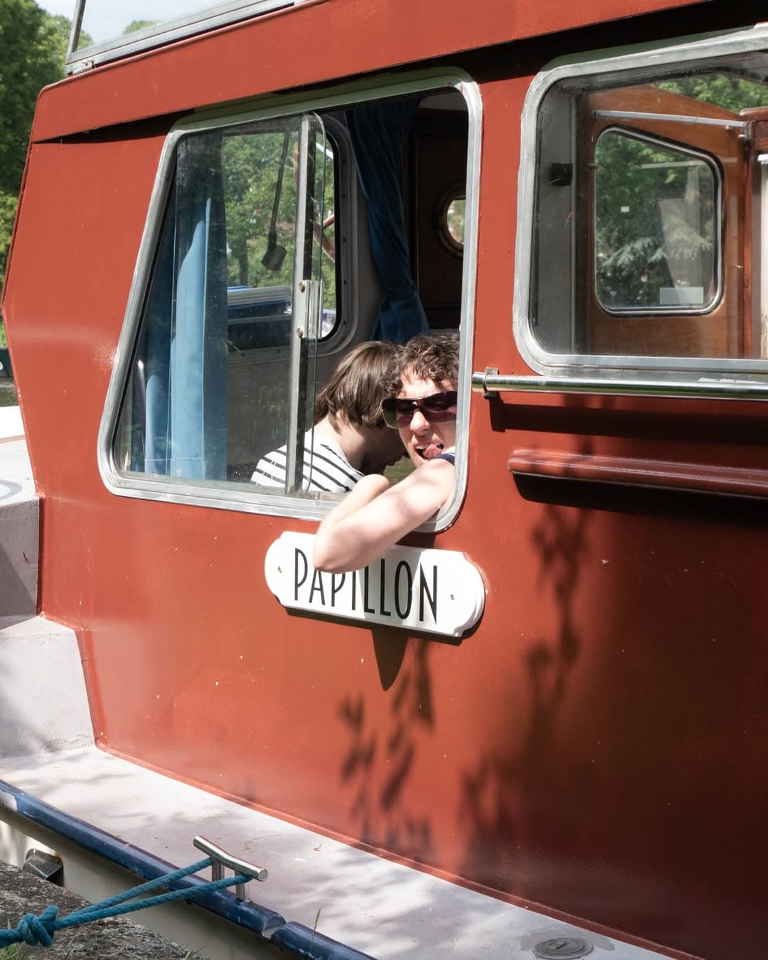 The image shows a maroon boat with someone looking out of the window sticking their tongue.