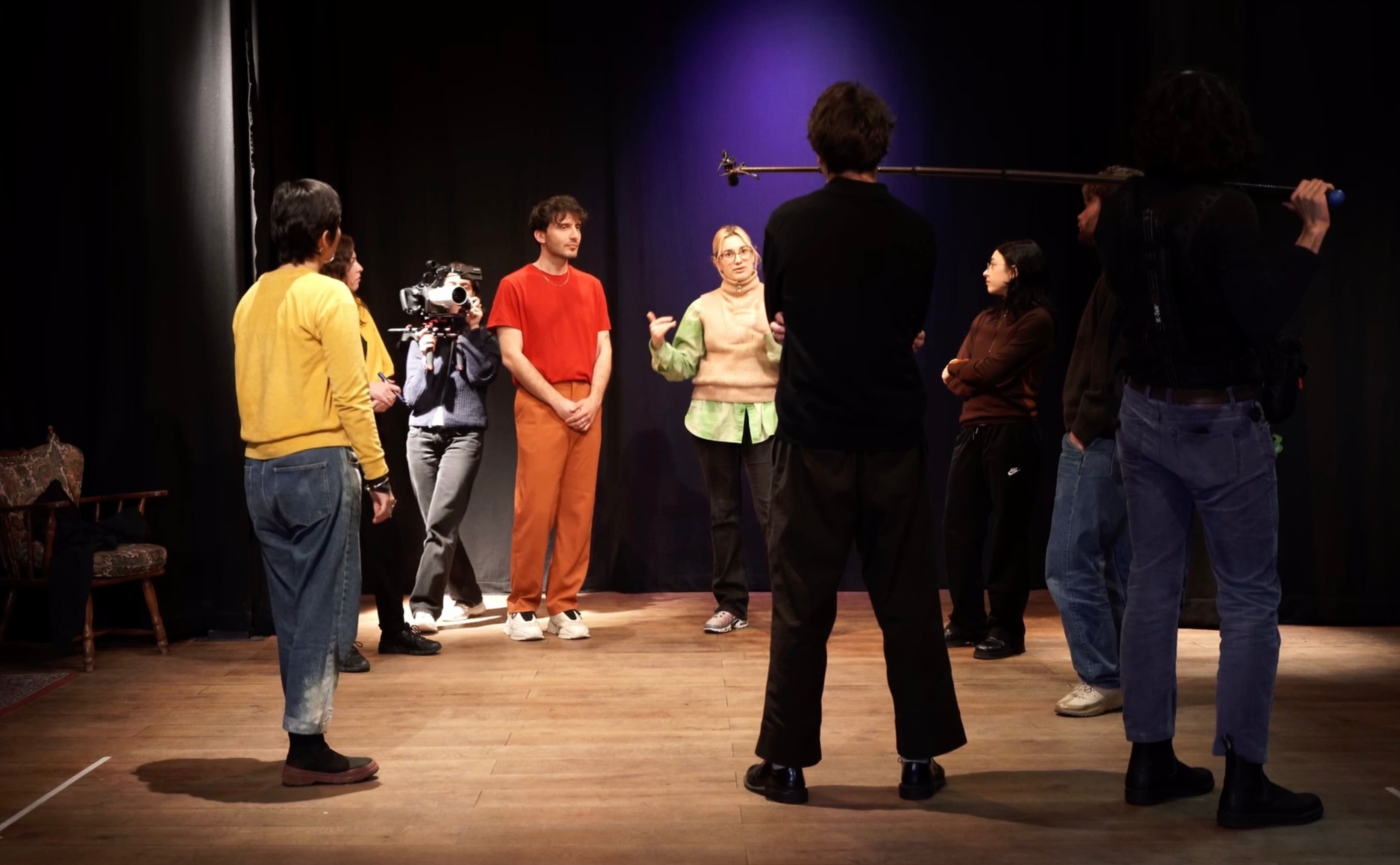 The image shows a group of people in a dark room, dimly lit, talking in a circle.
